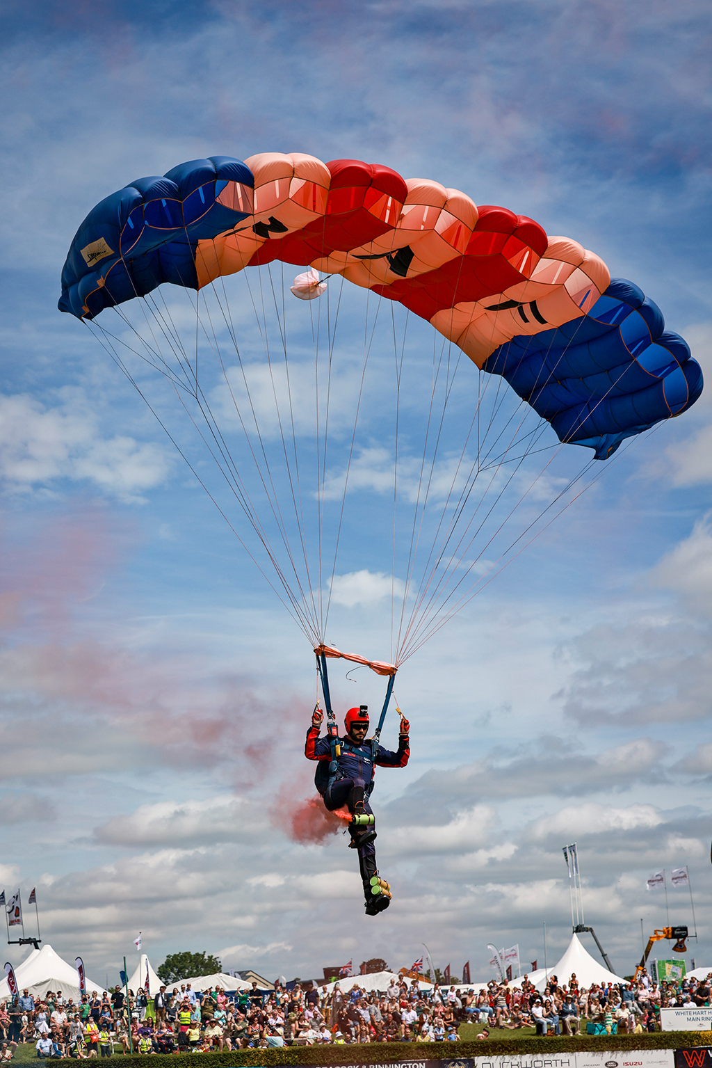 A lone RAF Falcon, wearing a red helmet and red trimmed dark blue jumpsuit descends under a red, white, and blue RAF parachute, trailing orange smoke above a large crowd at an outdoor airshow is about to land.